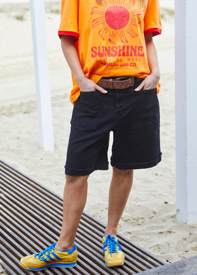 Hammill & Co pair of longer-length black stretch denim shorts with a relaxed fit. They have front pockets, a raw rolled hem, and sit mid-thigh