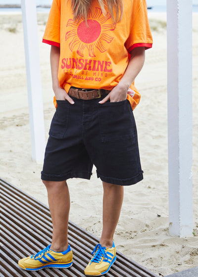 Hammill & Co pair of longer-length black stretch denim shorts with a relaxed fit. They have front pockets, a raw rolled hem, and sit mid-thigh